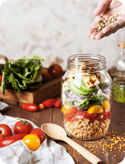 Bocal en verre rempli de salade composée (légumes frais, tomates cerises, graines et fromage), avec une main ajoutant des graines par-dessus, posé sur une table en bois entouré d’ingrédients frais.